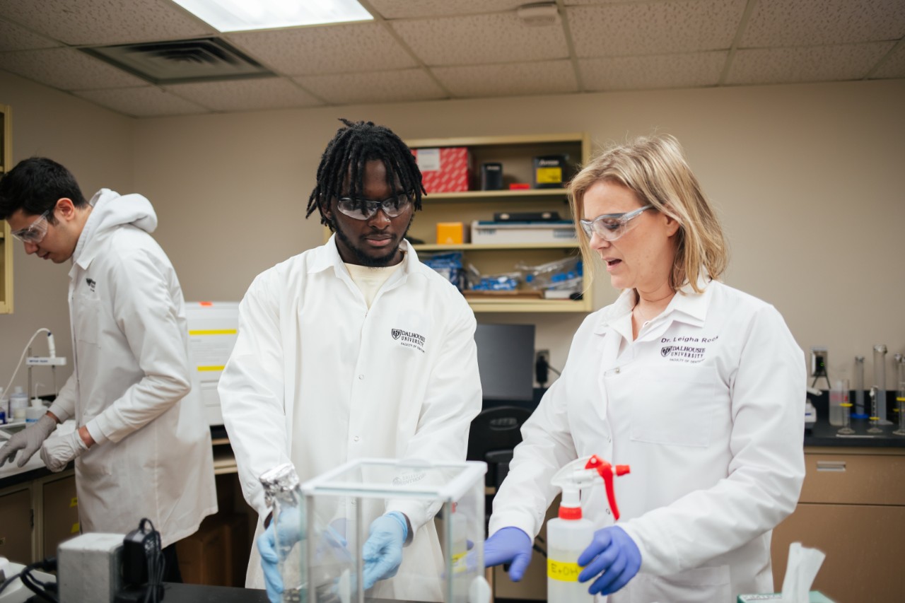 Three dalhousie researchers in a lab wearing lab coats and safety goggles.