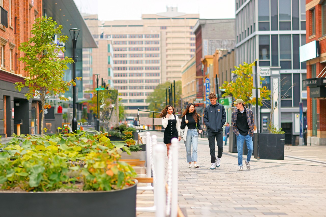 Four casually dressed students walk down a street paved in red bricks, with cityscape in background.