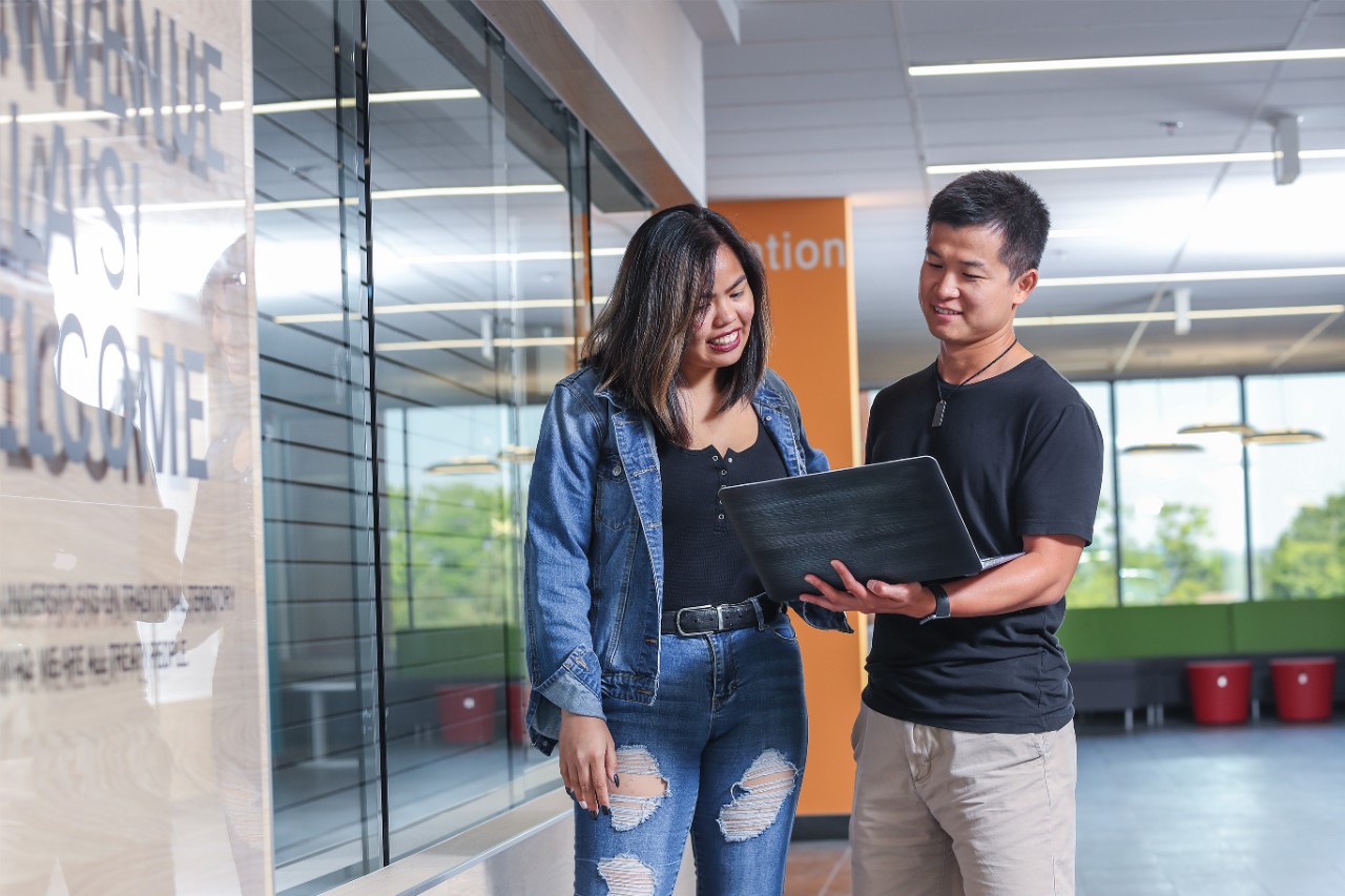 Male and female student stand in the library looking at an open laptop.